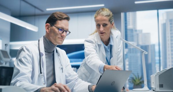 Two Doctors in Hospital Office Discuss a Diagnosis, Analyzing Data on a Laptop. Male and Female Medical Professionals Talking about Treatment, Drug Prescription during Telemedicine Call