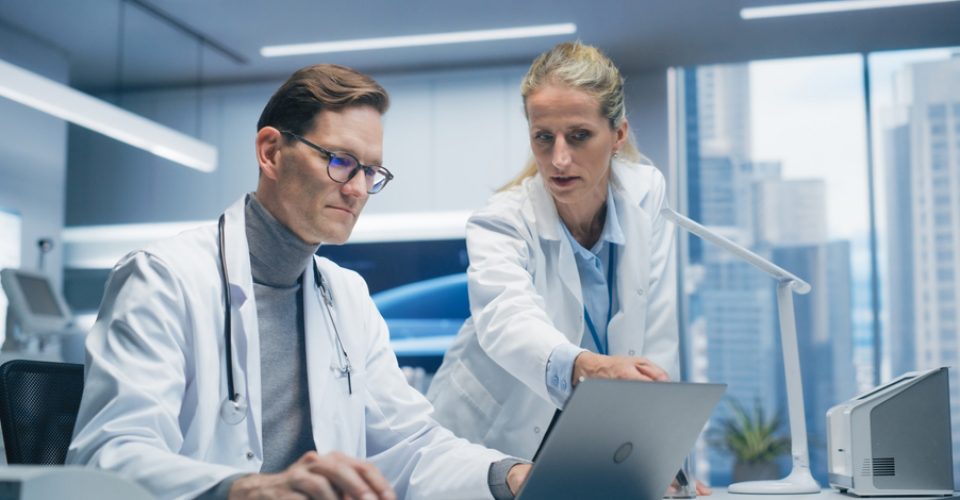 Two Doctors in Hospital Office Discuss a Diagnosis, Analyzing Data on a Laptop. Male and Female Medical Professionals Talking about Treatment, Drug Prescription during Telemedicine Call